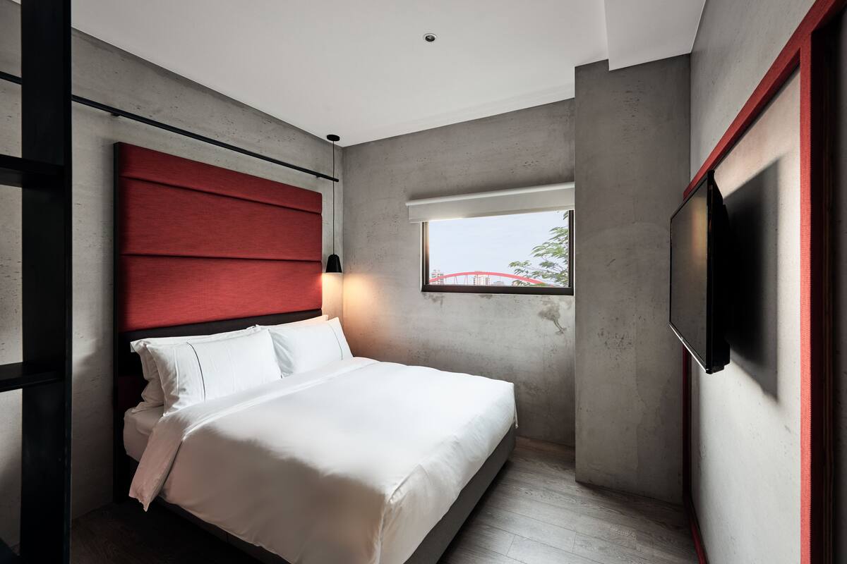 Un dormitorio moderno con cama, cabecera roja, ventana con vista a un puente y televisor montado.