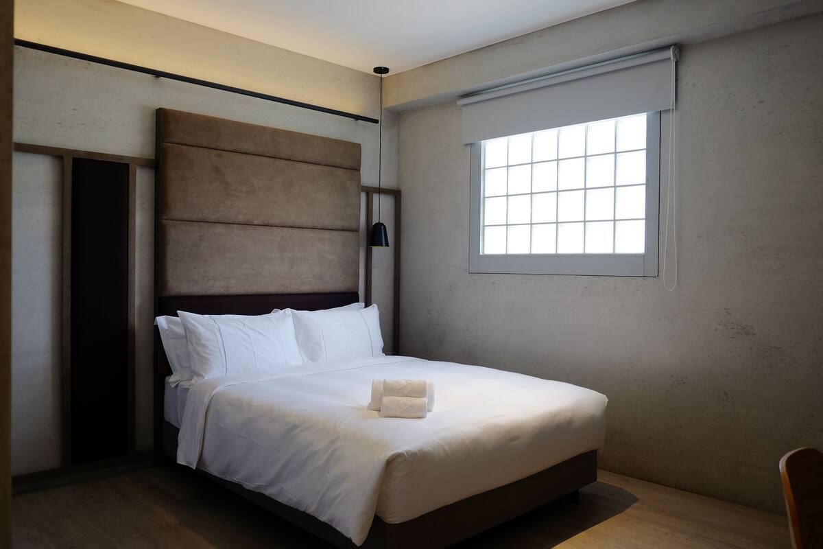 Una cama bien hecha con sábanas blancas y una toalla enrollada, un cabecero, una ventana con persianas y un suelo de madera.