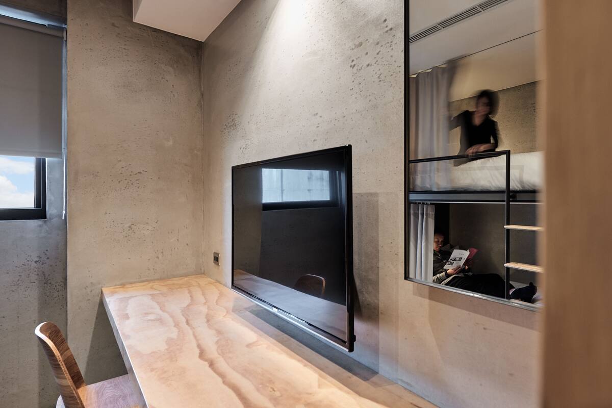 Habitación moderna con un televisor de pantalla plana grande montado en una pared de hormigón, una mesa de madera y una cama litera con una persona leyendo.