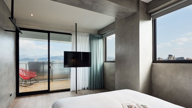 A modern hotel room with a large bed, a flat-screen TV, a balcony with a red chair, and a city view. - Waterfront Hotel (Taipei)