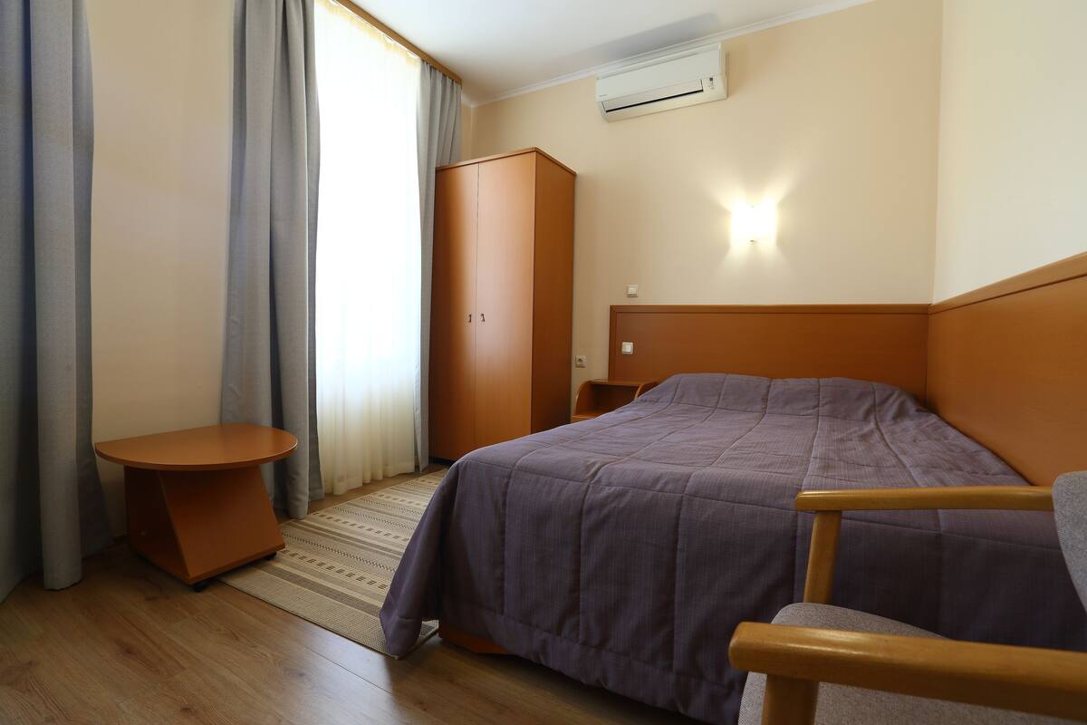 A hotel room with a bed, a wooden chair, a small table, a wardrobe, and an air conditioning unit.