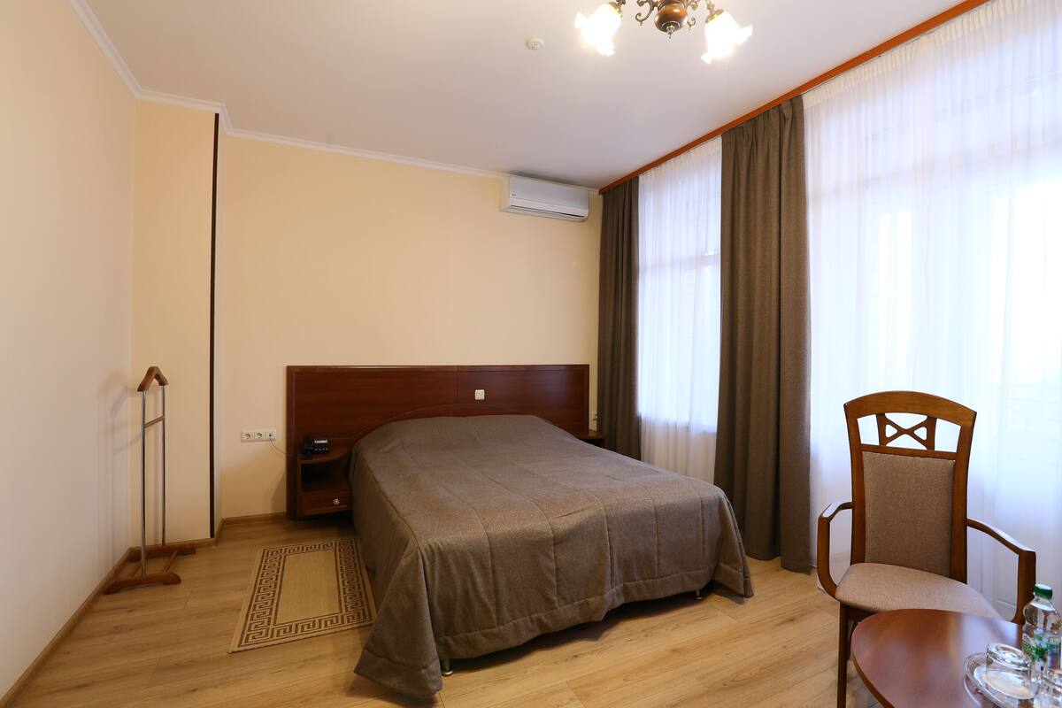 A hotel room with a large bed, a wooden chair, a small table, and a standing heater.