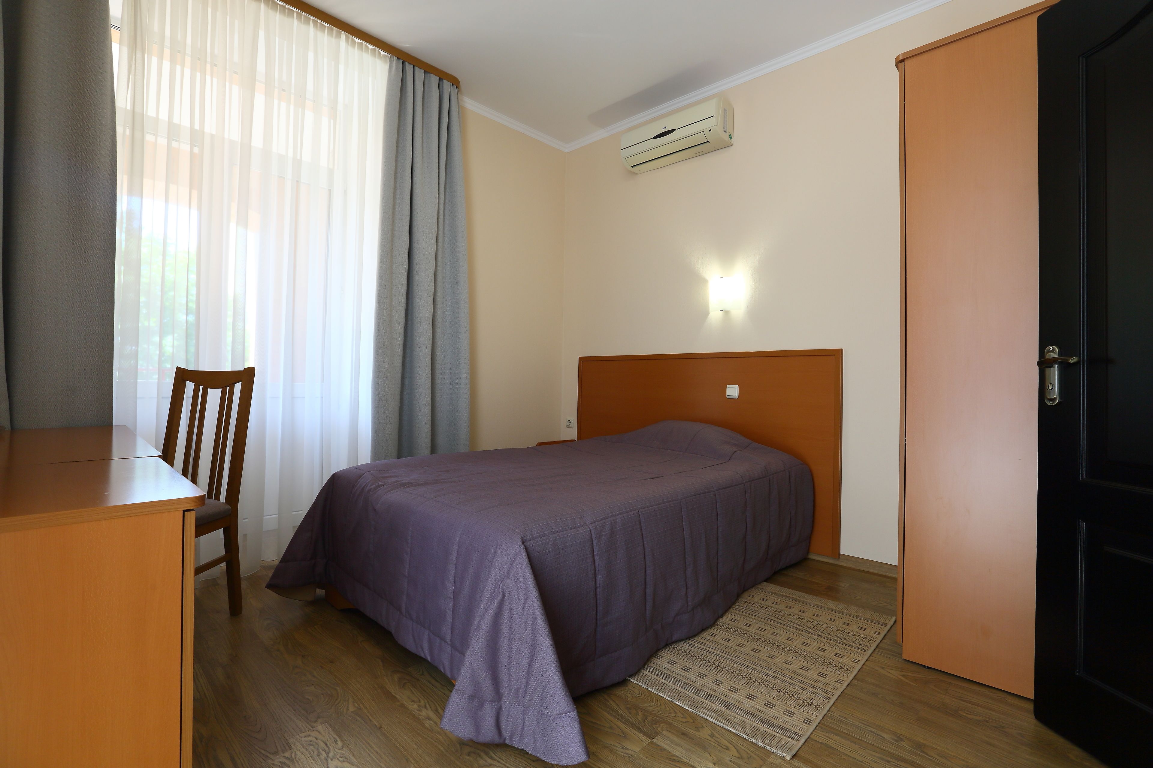 Un dormitorio con cama, escritorio, silla, puerta y ventana con cortinas.