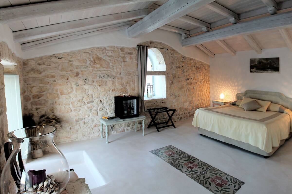 A bedroom with a stone wall, a bed, a nightstand, a table with a lamp, and a window.