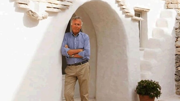 A man standing in a white-walled corridor with a stone wall and a potted plant. - Palazzo Camarda (Ceglie Messapica)