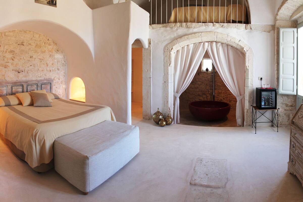 A bedroom with a large bed, a stone staircase, a red bathtub, and a small wooden dresser.