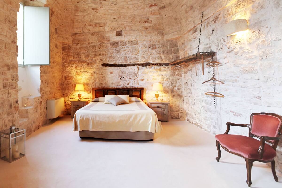 A bedroom with a stone wall, a bed with two bedside tables, a hanging light fixture, and a red chair.