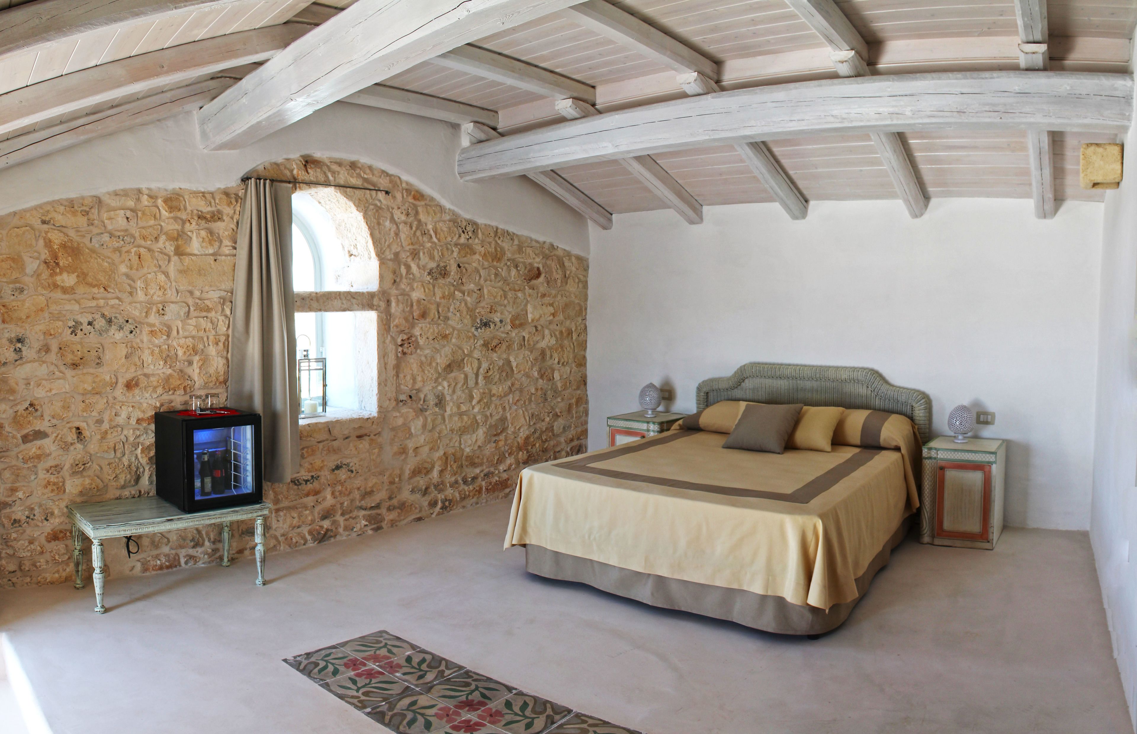Une chambre avec un mur en pierre, un lit recouvert d’une couverture beige et marron, une petite table avec un téléviseur et une fenêtre avec des rideaux.