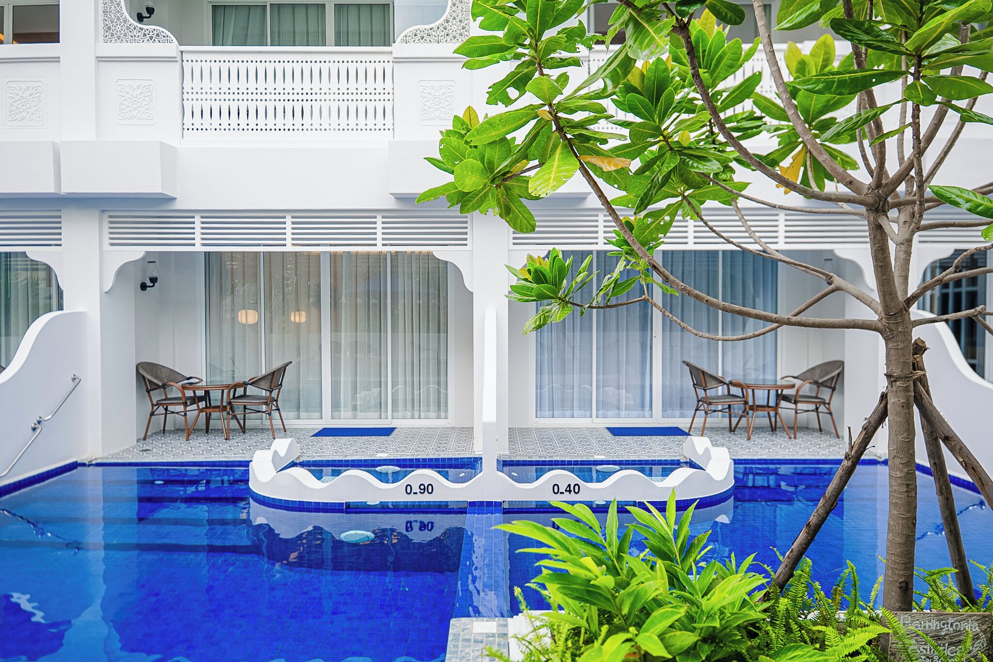 A pool area with two seating areas, each with a table and chairs, surrounded by lush greenery.