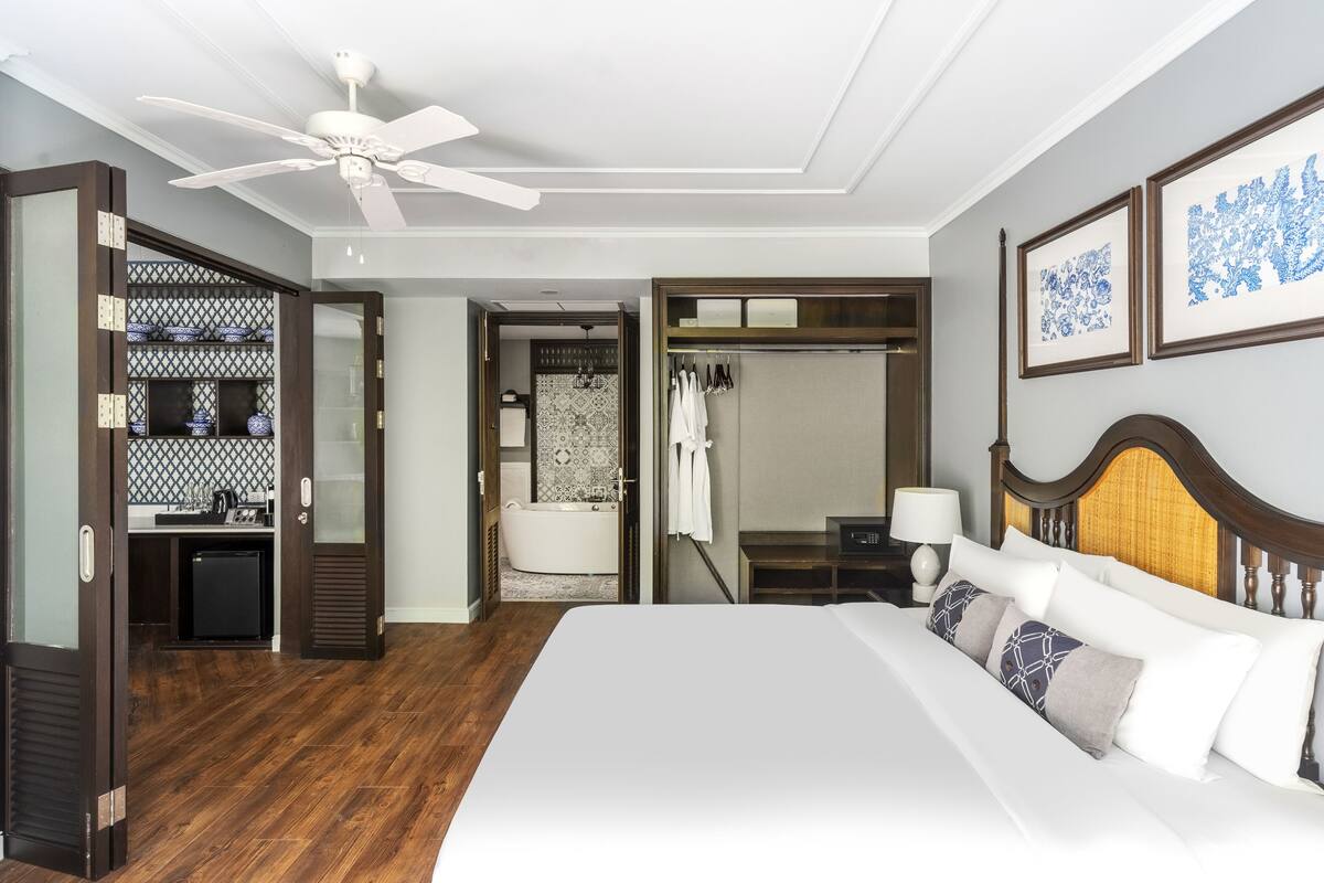 Ein Hotelzimmer mit einem großen Bett, einem Deckenventilator, einem Kleiderschrank, einem Badezimmer mit Badewanne sowie einem kleinen Schreibtisch mit Lampe.