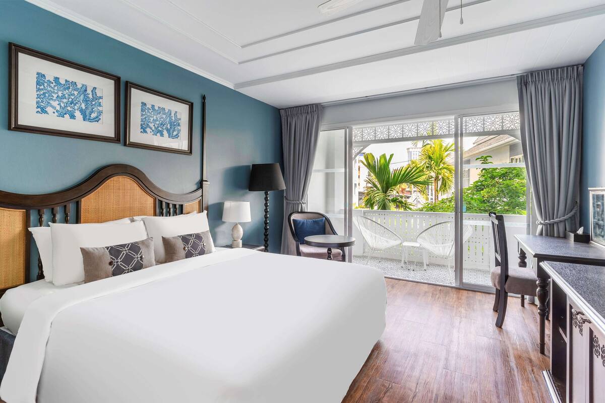 Ein Hotelzimmer mit einem großen Bett, einem Schreibtisch, einem Stuhl und einem Balkon mit Blick auf Palmen.