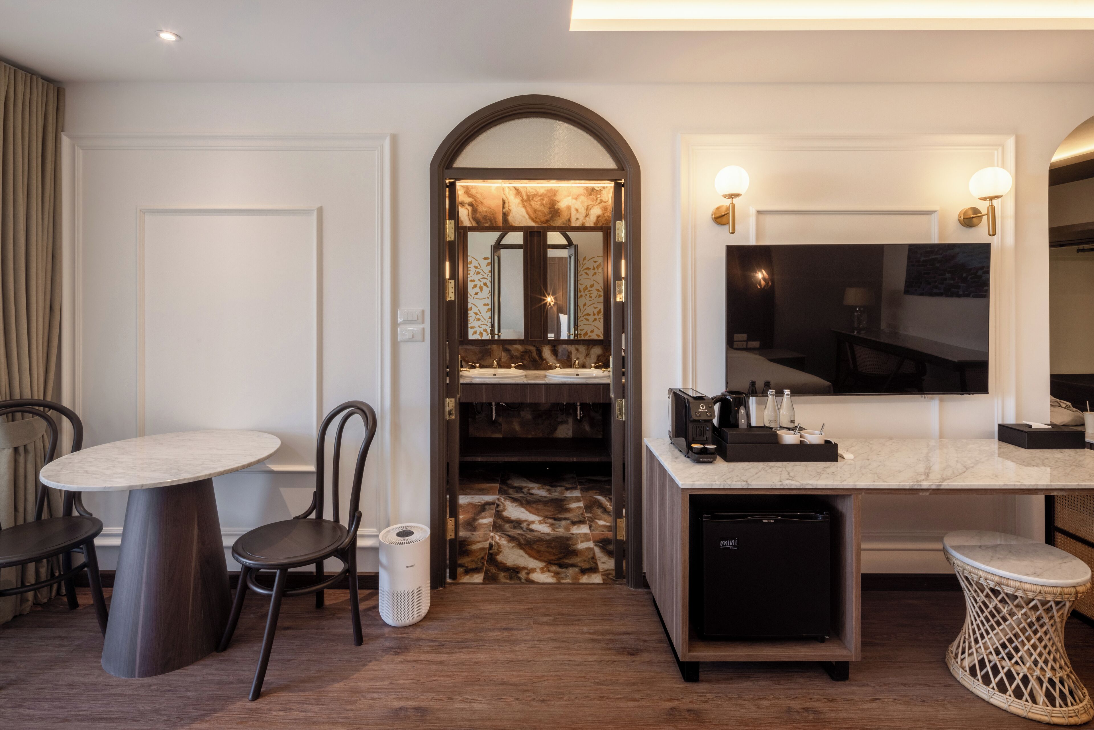 A compact hotel room with a dining area, a small kitchen with a refrigerator, a bathroom with a vanity, and a large mirror.