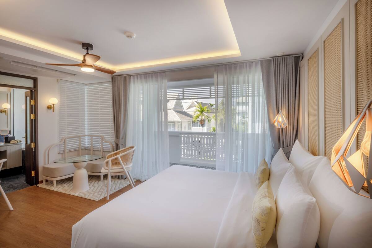 Ein modernes Hotelzimmer mit einem großen Bett, einem Glastisch und einem Stuhl, einem Deckenventilator und Blick auf den Balkon.