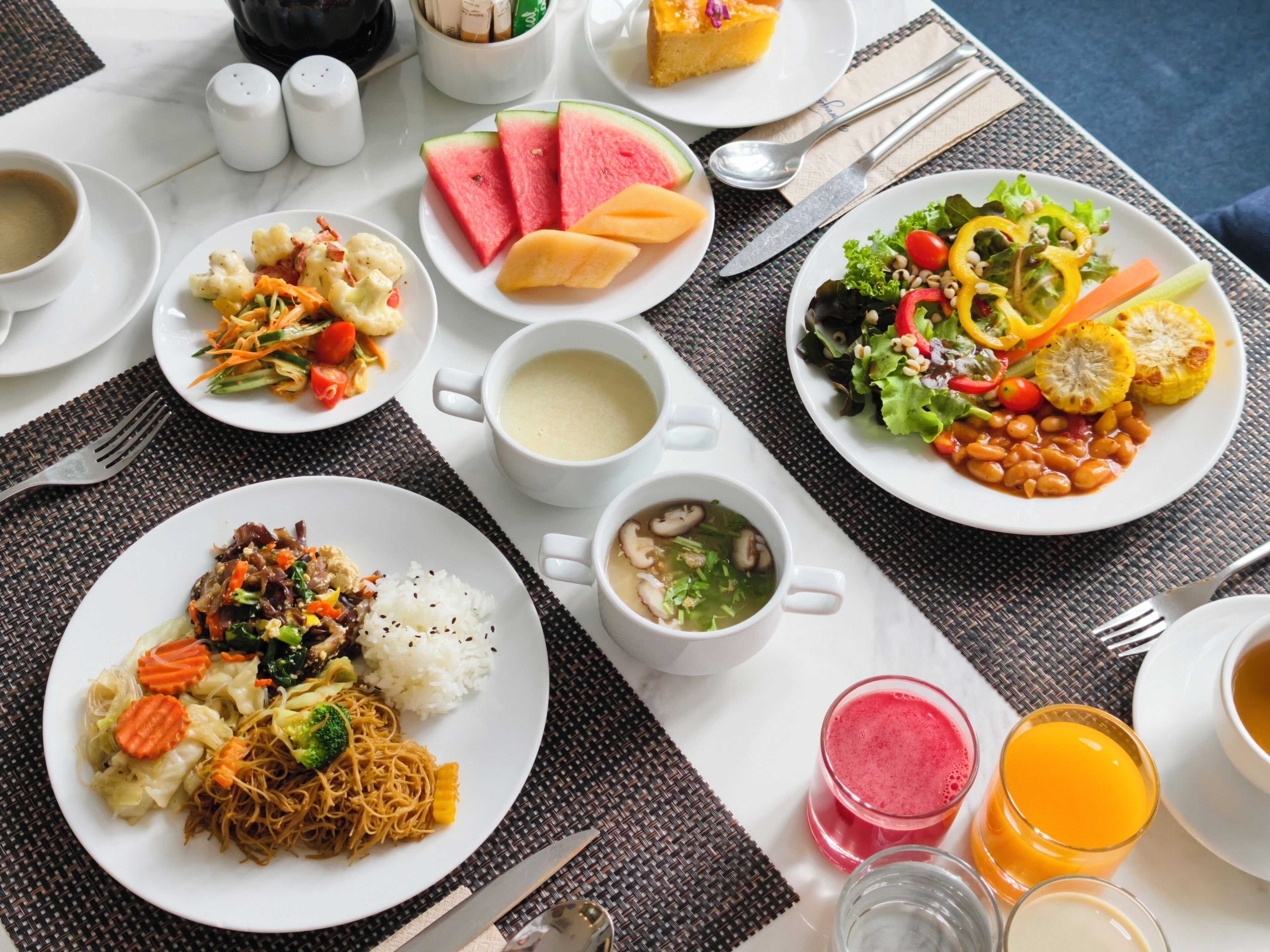 A table set with various dishes, including rice, noodles, soup, and a salad, along with drinks and condiments.