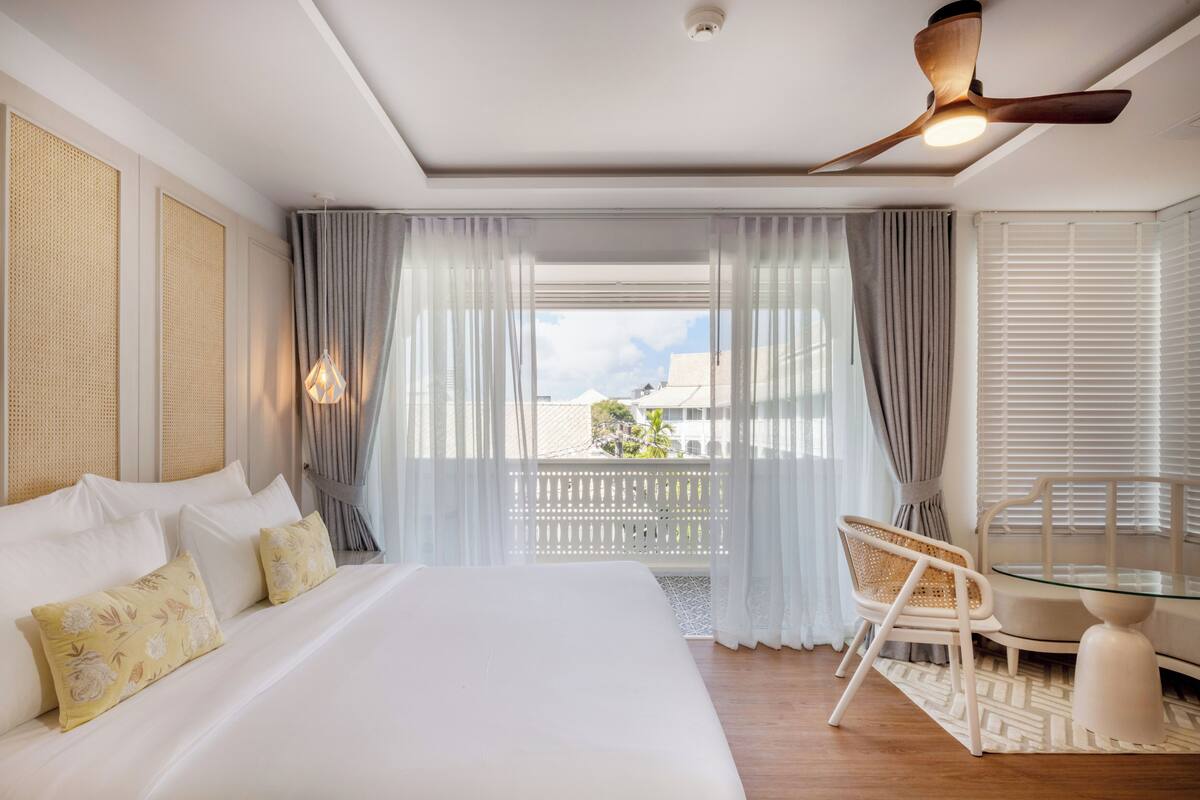 Ein Hotelzimmer mit einem großen Bett, einem Glastisch, einem Stuhl und einem Balkon mit Vorhängen.