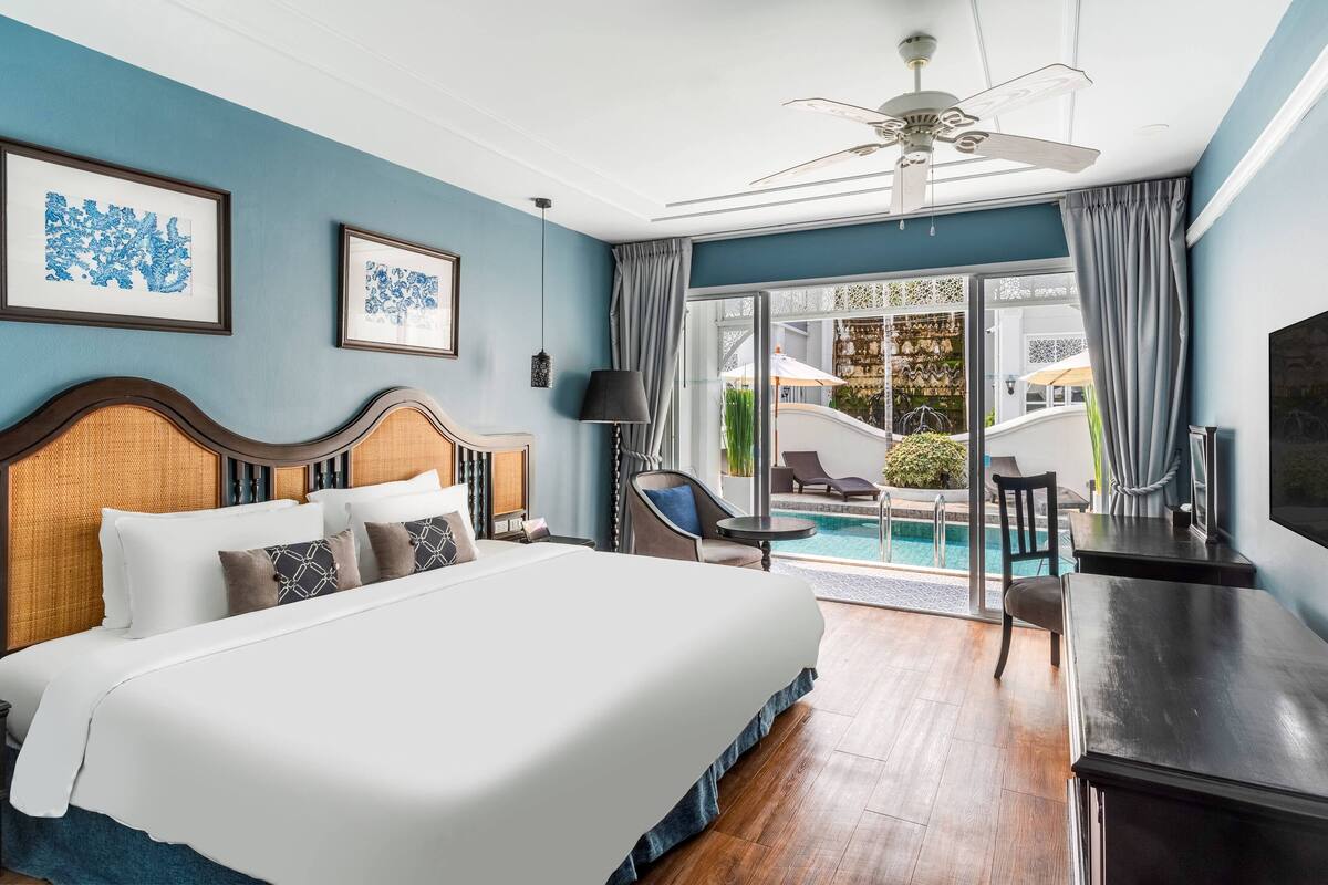 Ein Hotelzimmer mit einem großen Bett, einem Deckenventilator, einem Schreibtisch und Blick auf einen Außenpool.
