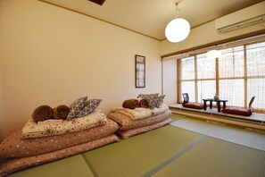 A tatami mat room with a futon bed, a small table with a vase, and a window with bamboo blinds.