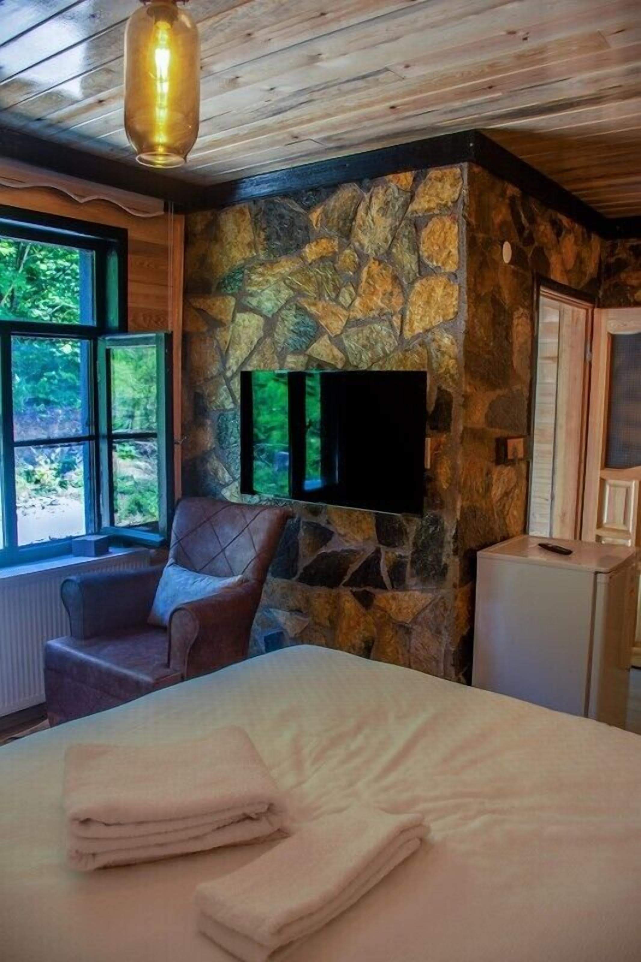 A room with a stone wall, a bed with folded towels, a leather chair, a television mounted on the wall, and a window with a view of greenery.