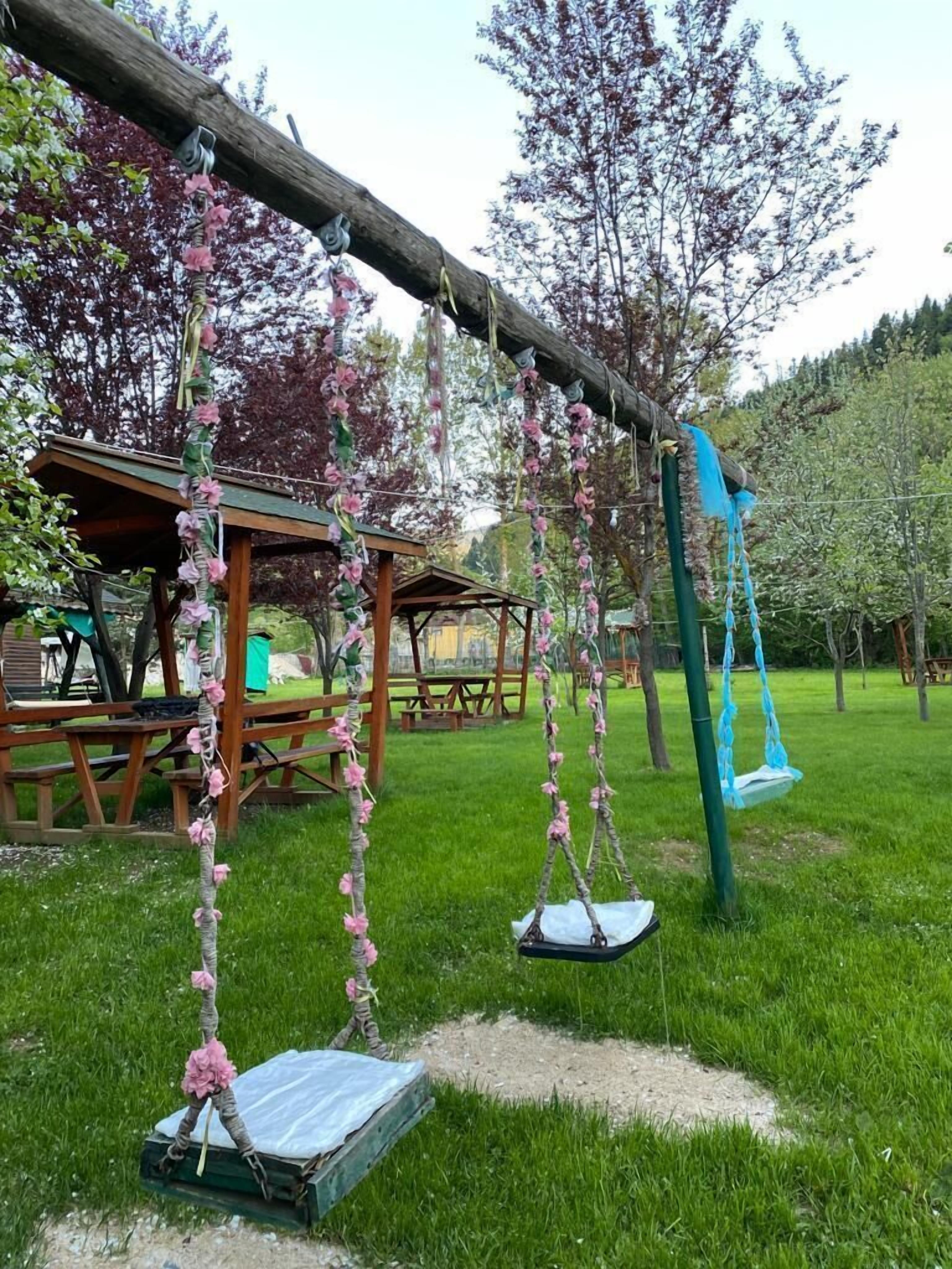 Un parc infantil de fusta amb un cadira d'oscil·lació blava, envoltat d'herba verda i arbres amb flors rosades.