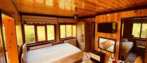 A wooden cabin interior with a bed, a sofa, a television, and a window with a view of greenery.