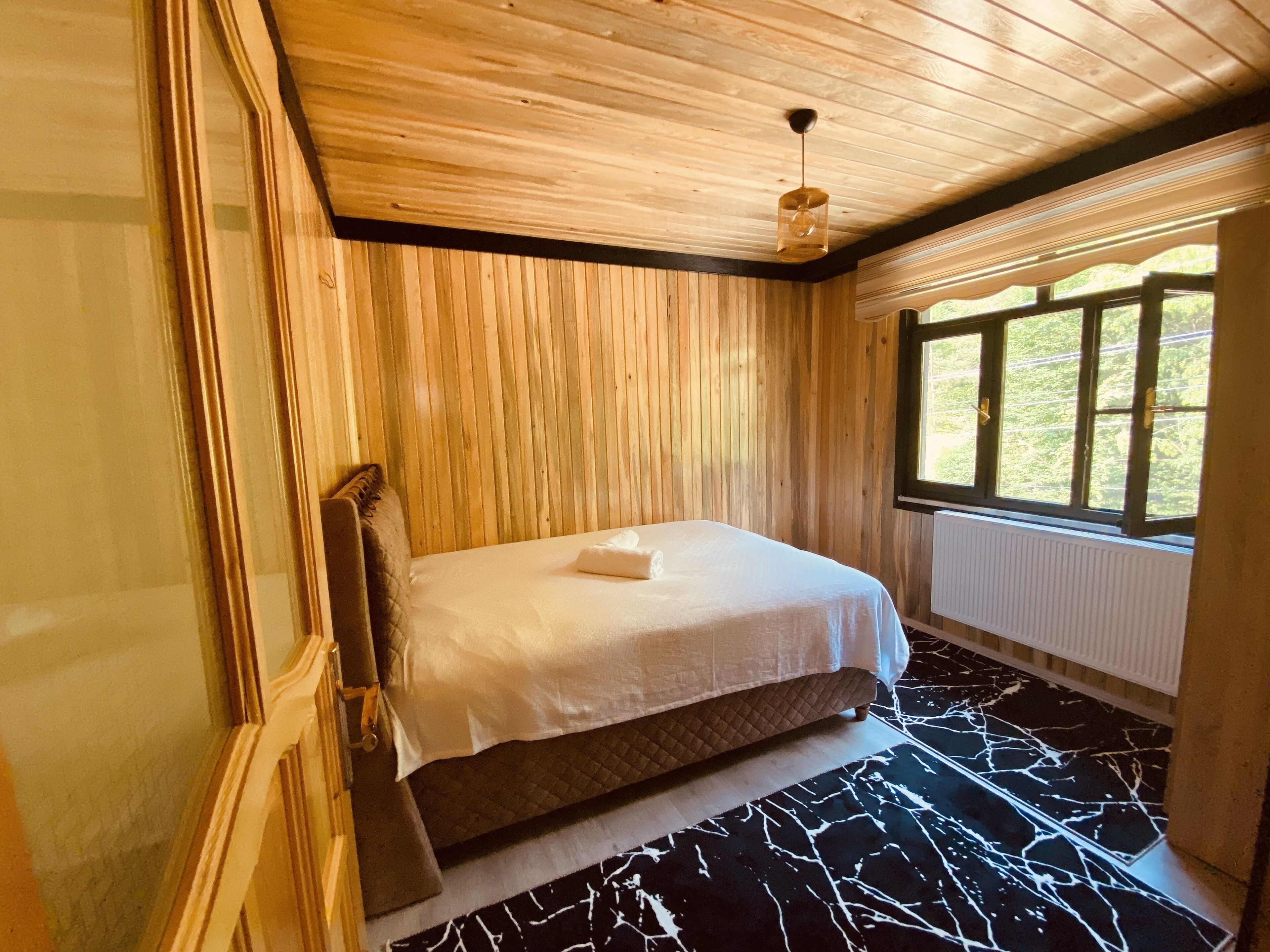 A bedroom with a wooden ceiling, a bed with a quilted headboard, a window with a view of greenery, and a patterned floor.