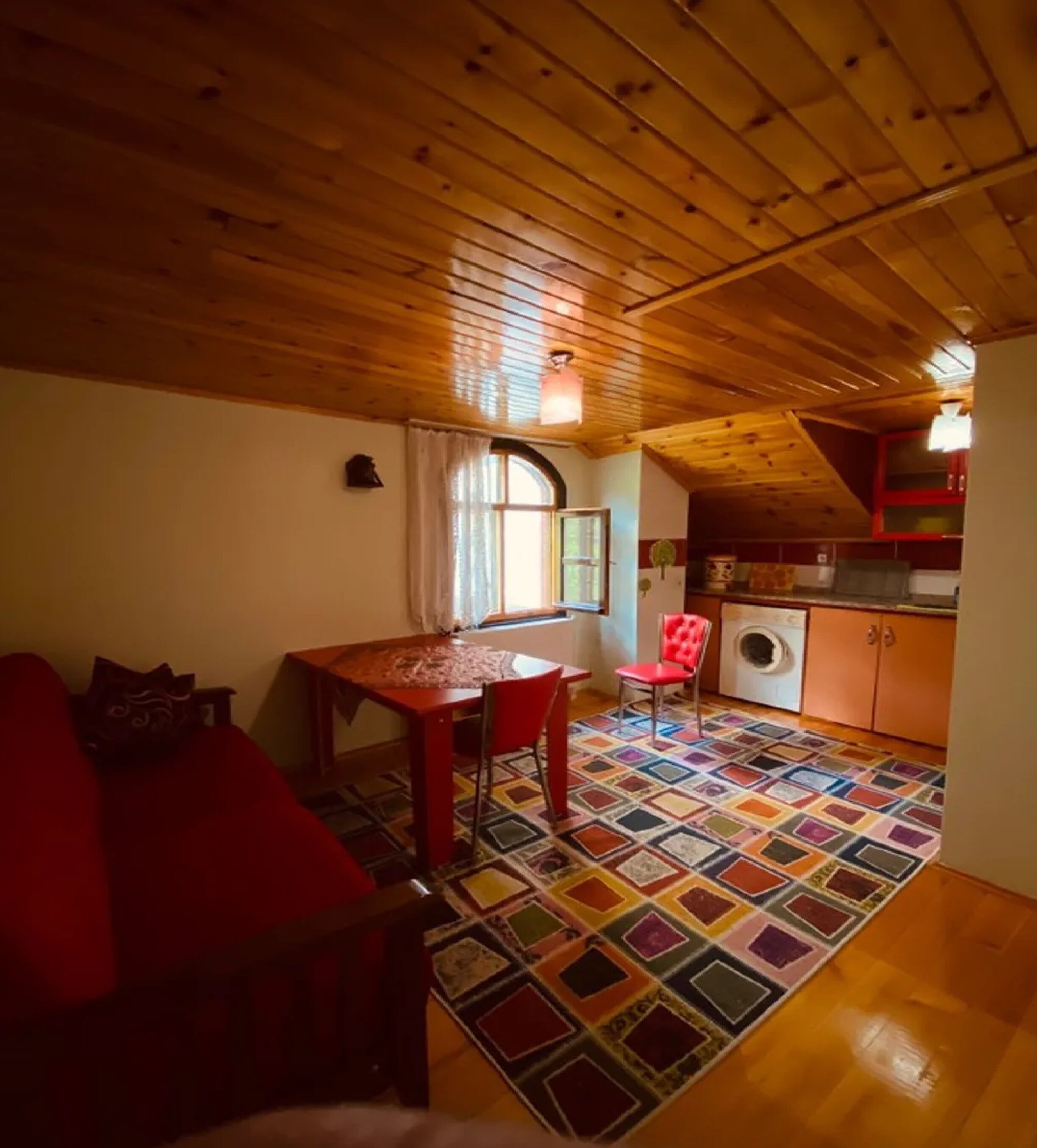 A cozy room with a wooden ceiling, a dining table, a red sofa, a colorful rug, and a kitchen area with a washing machine.