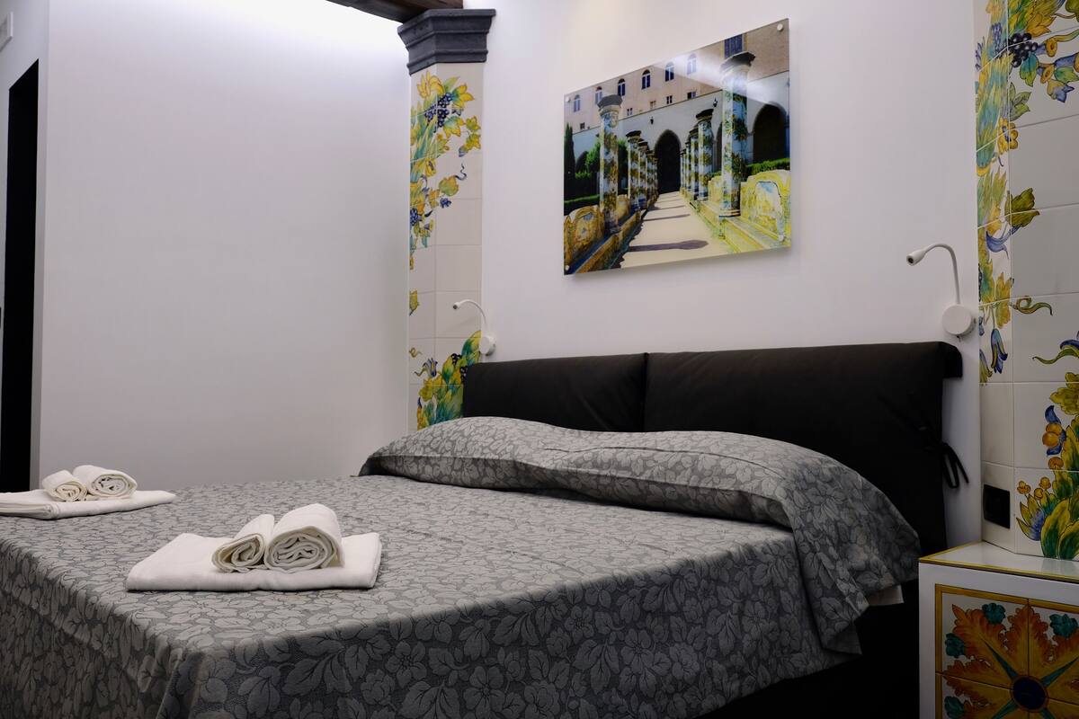A hotel room with a bed, a painting of a street scene, and floral-patterned walls.