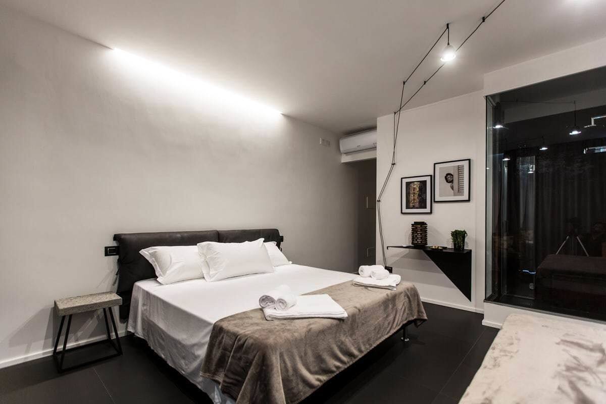 A modern bedroom with a large bed, a small bench, a bedside table with a lamp, and a large window with a view.