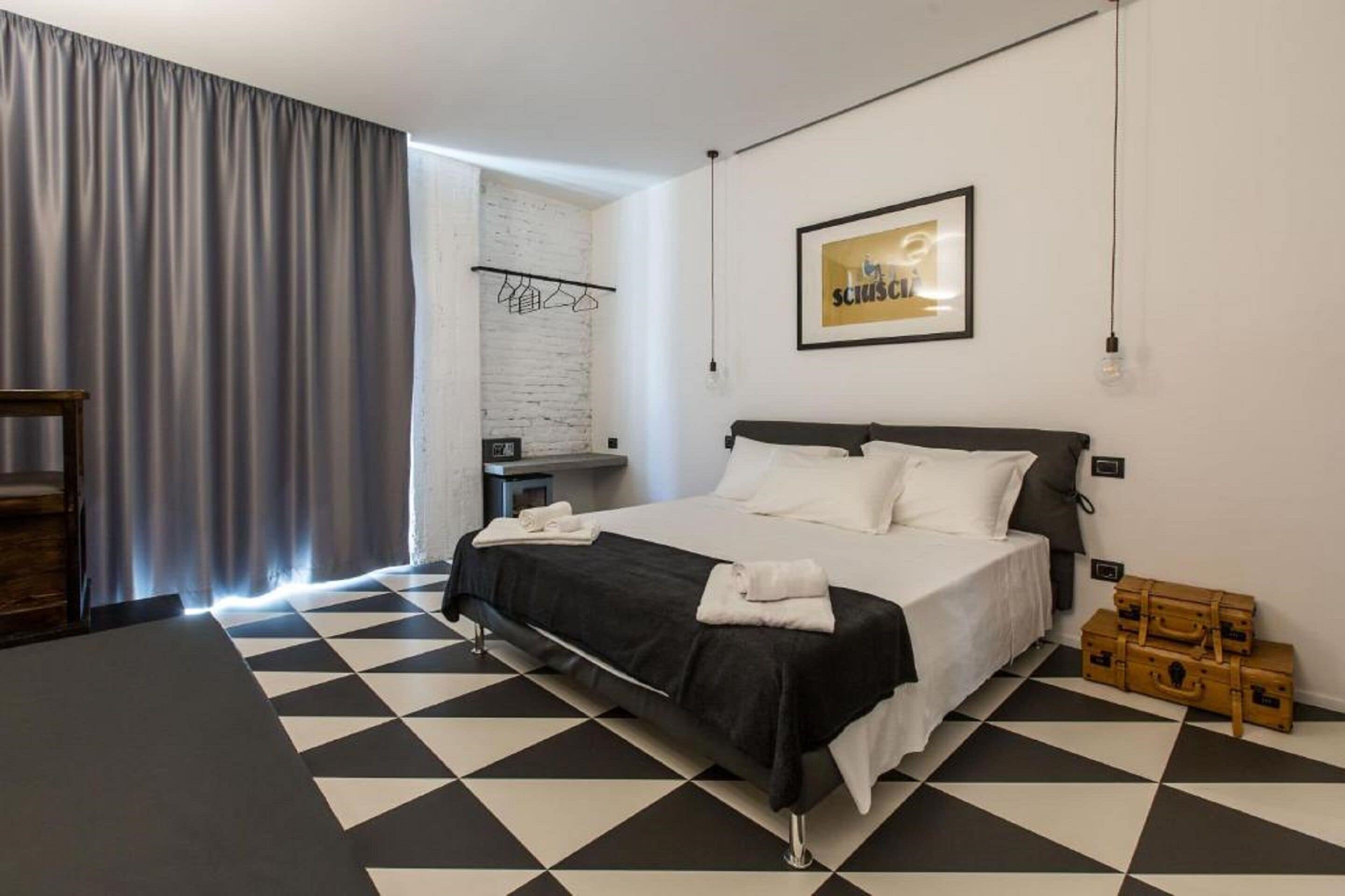 A modern hotel room with a black and white checkered floor, a bed with white linens, a grey curtain, a wall-mounted light fixture, and a framed picture on the wall.