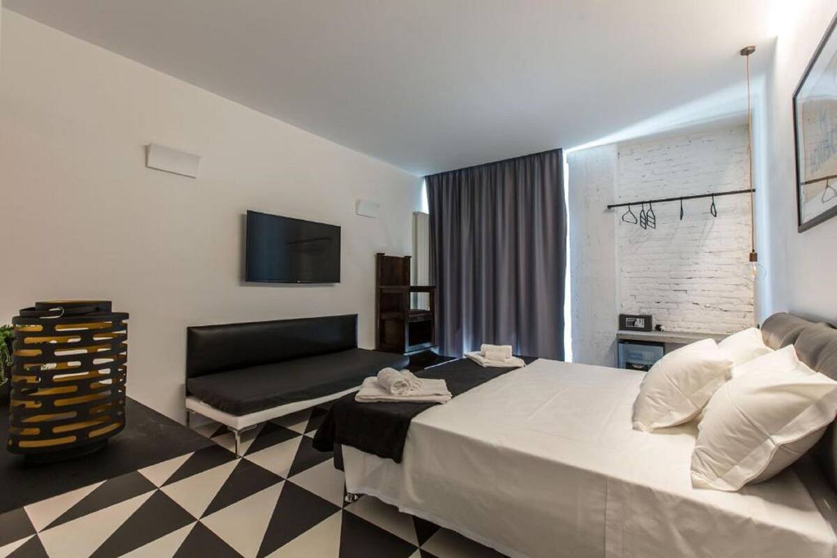 A modern hotel room with a black and white checkered floor, a bed with white linens, a black sofa, a television mounted on the wall, and a white brick accent wall.