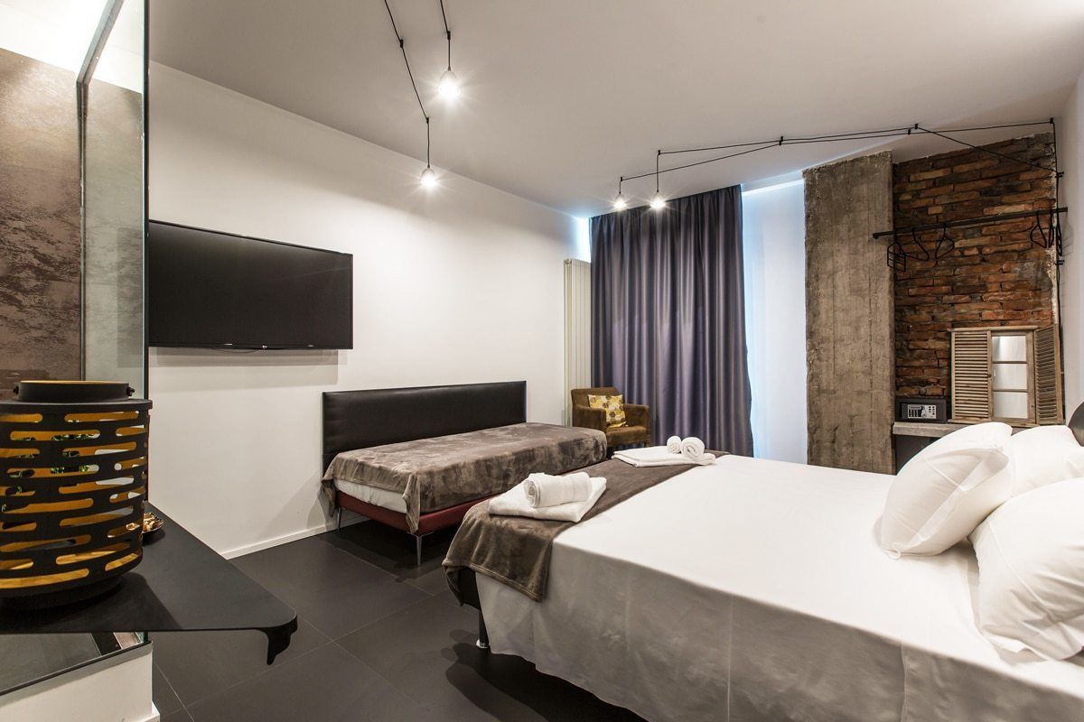 A modern hotel room with a large bed, a smaller bed, a flat-screen TV mounted on the wall, a stone accent wall, and a large window with curtains.