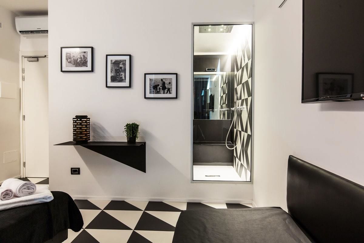Une chambre d’hôtel moderne avec un sol à carreaux noirs et blancs, un lit doté d’une tête de lit noire, une petite étagère ornée d’objets décoratifs et un téléviseur fixé au mur.