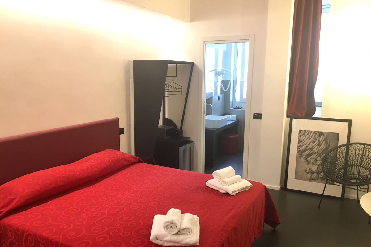 A hotel room with a red bedspread, a desk with a mirror, a framed picture, and a chair.