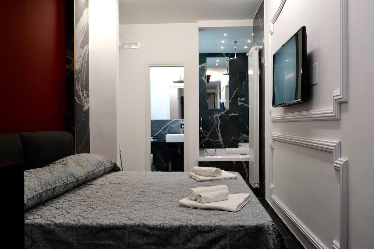A modern hotel room with a bed, a TV mounted on the wall, and a bathroom visible through an open door.