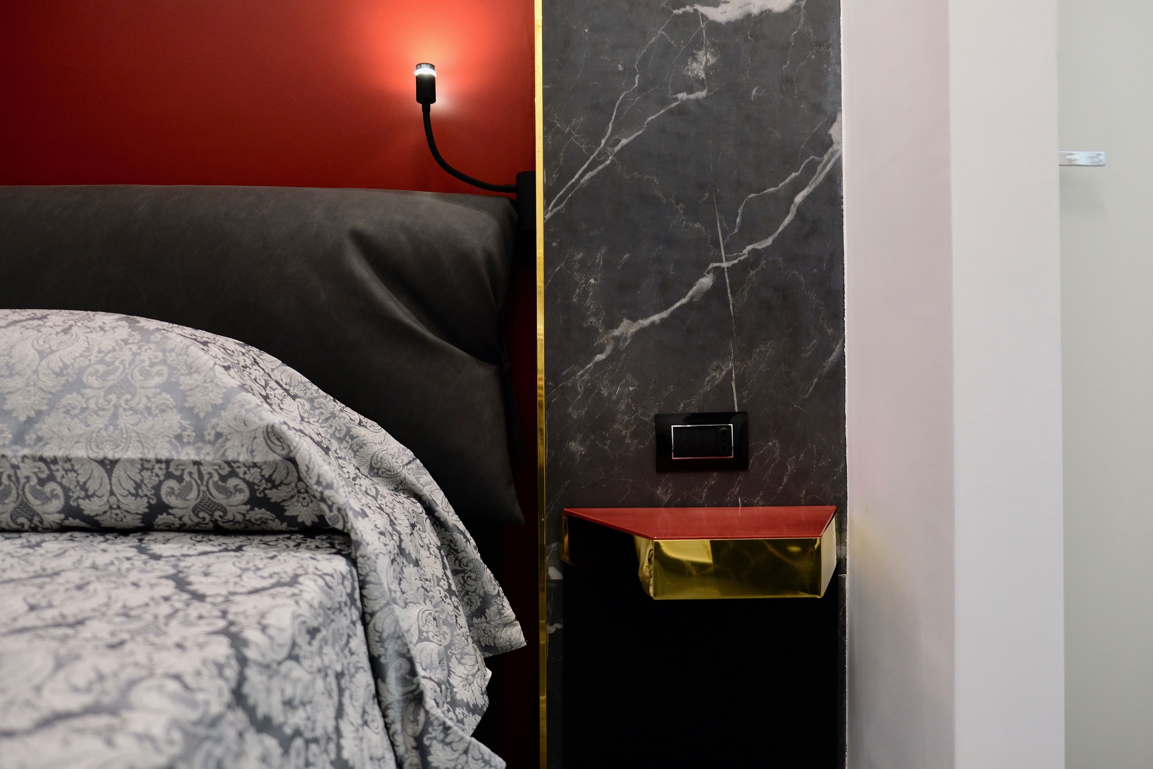 A bed with patterned bedding, a bedside table with a red top, and a wall-mounted light.