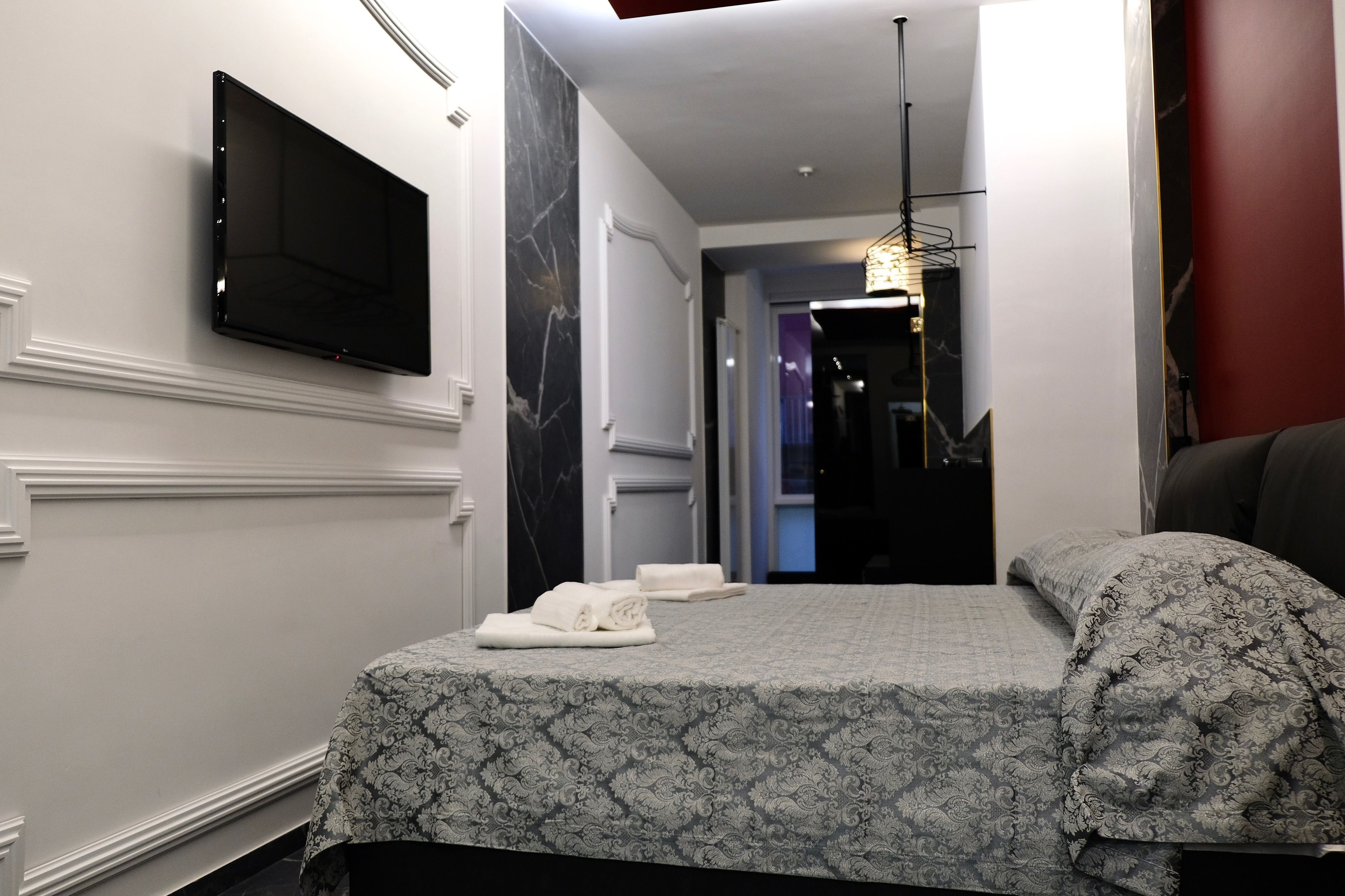 A hotel room with a bed, a flat-screen TV mounted on the wall, and a hallway leading to other rooms.