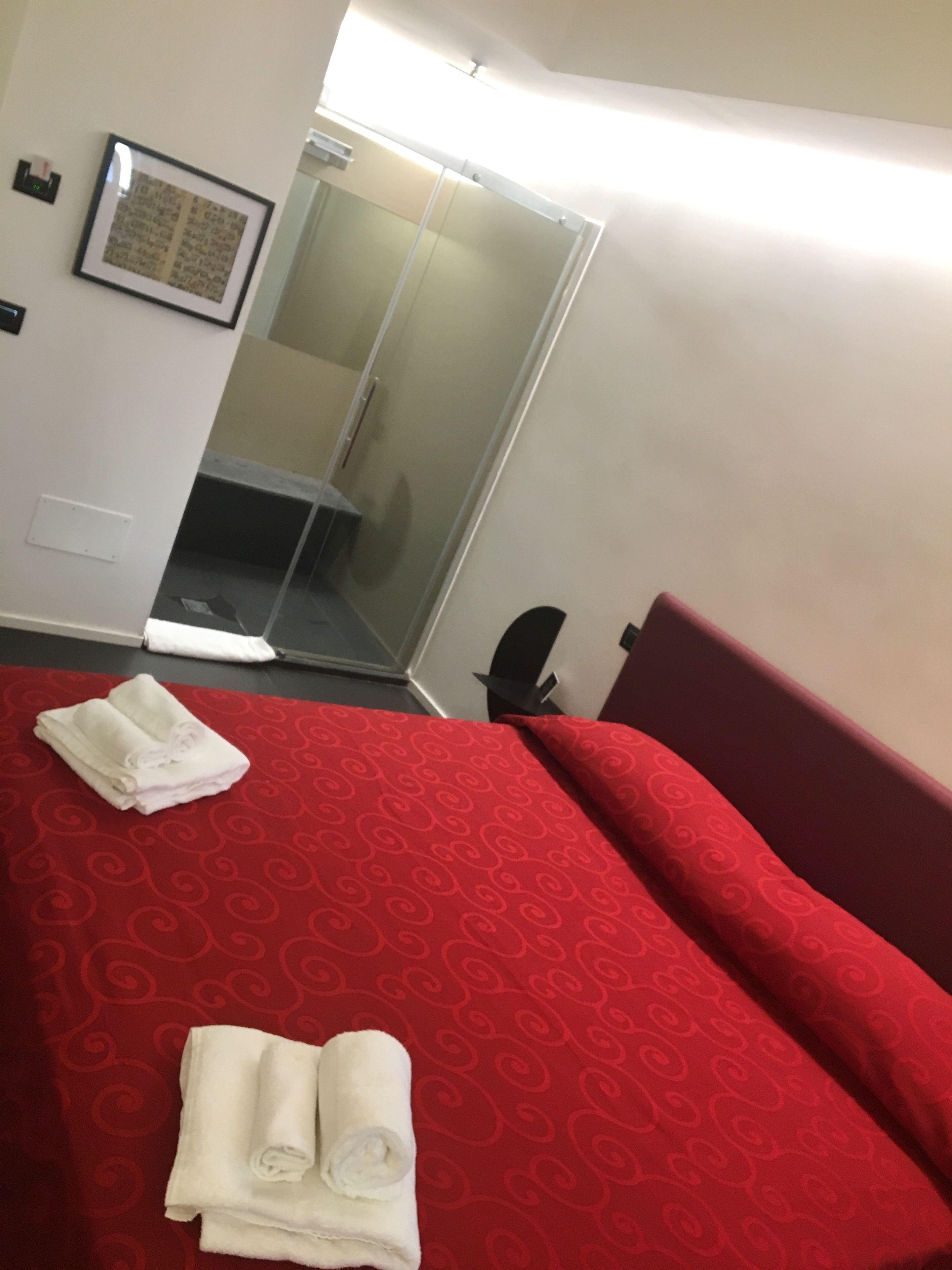 A hotel room with a bed, a red patterned bedspread, two rolled towels, a mirror, and a door leading to a bathroom.