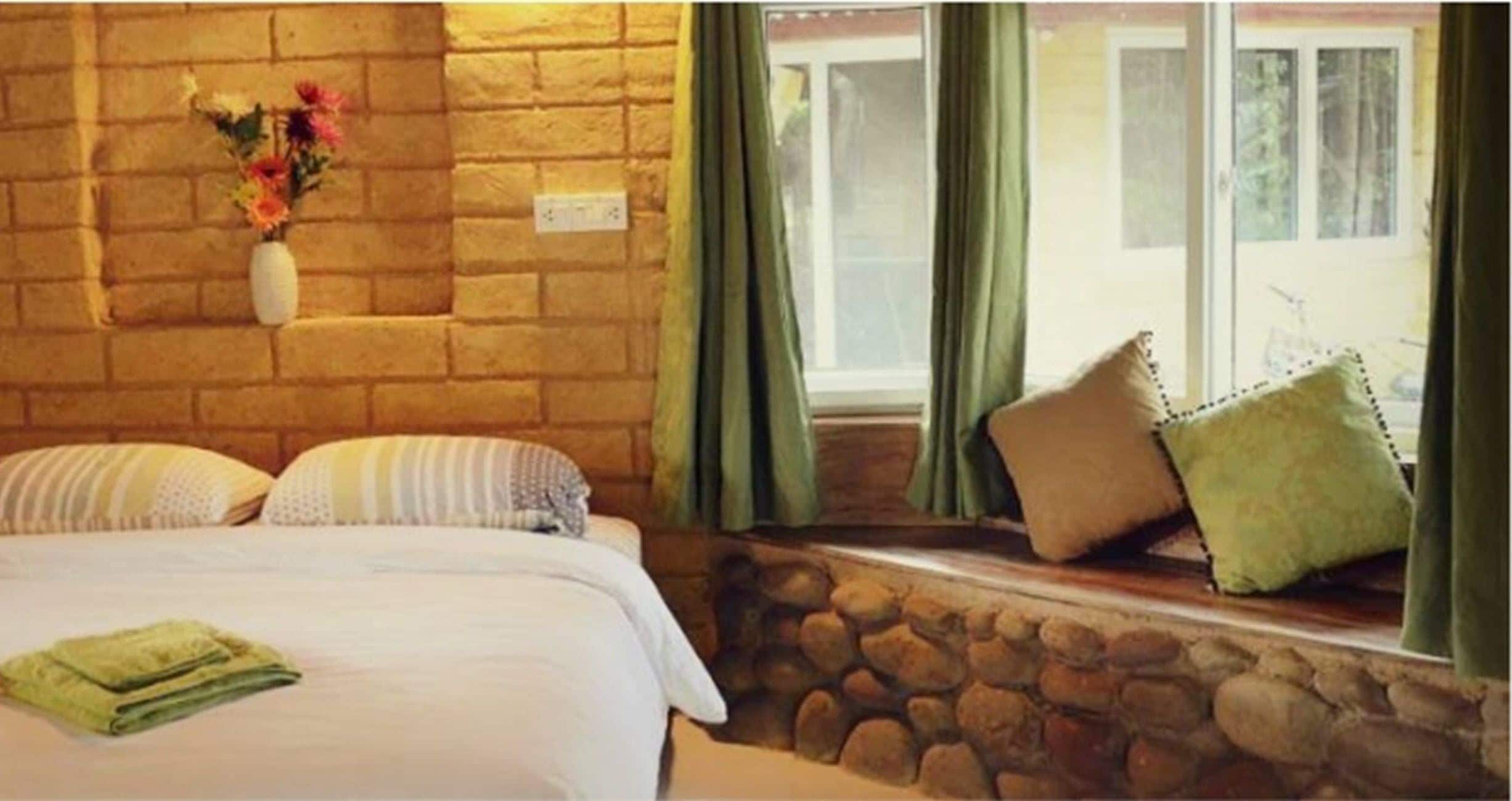 A bedroom with a stone wall, a bed with white bedding, a vase with flowers, and a window with curtains.