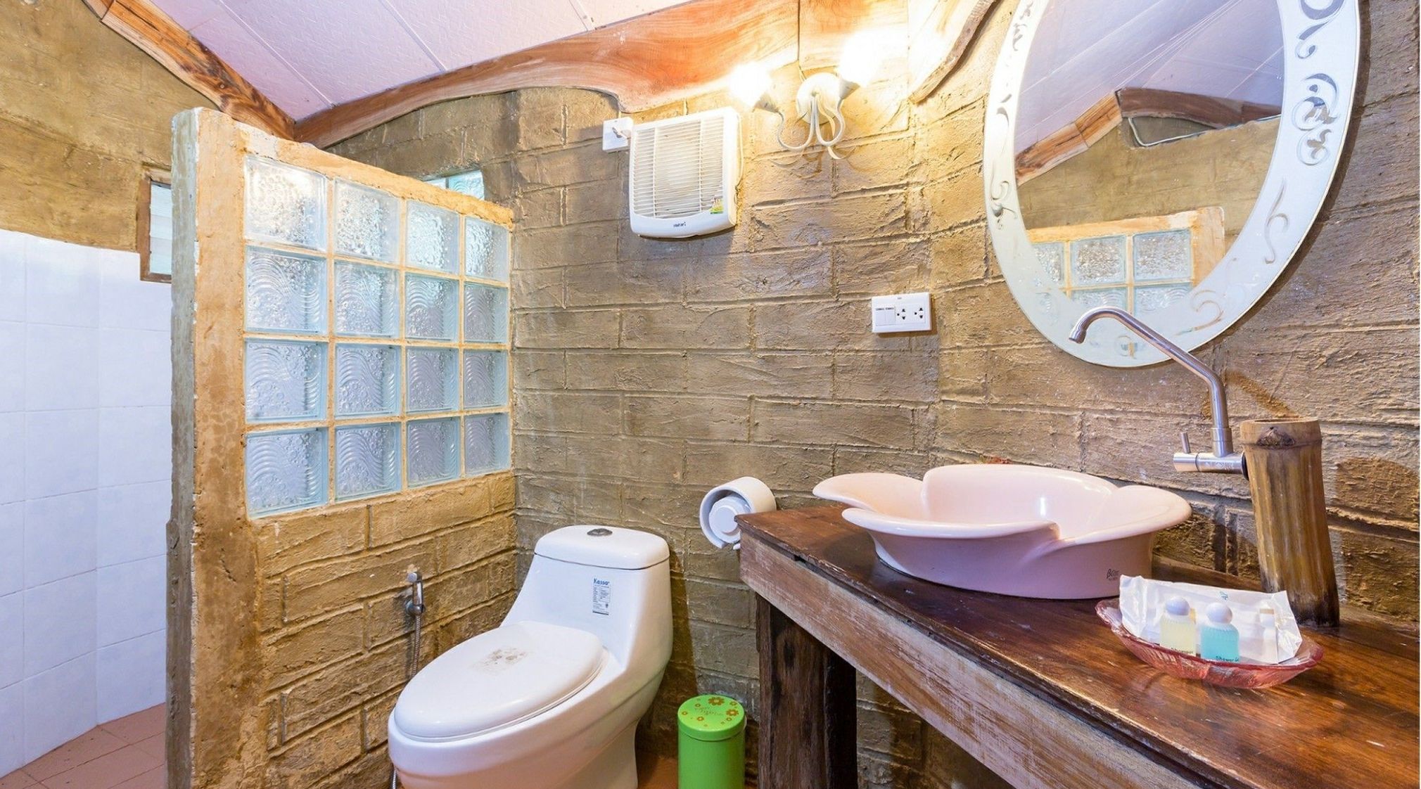Un baño rústico con pared de piedra, inodoro blanco, cubo de basura verde y tocador de madera con un lavabo grande.