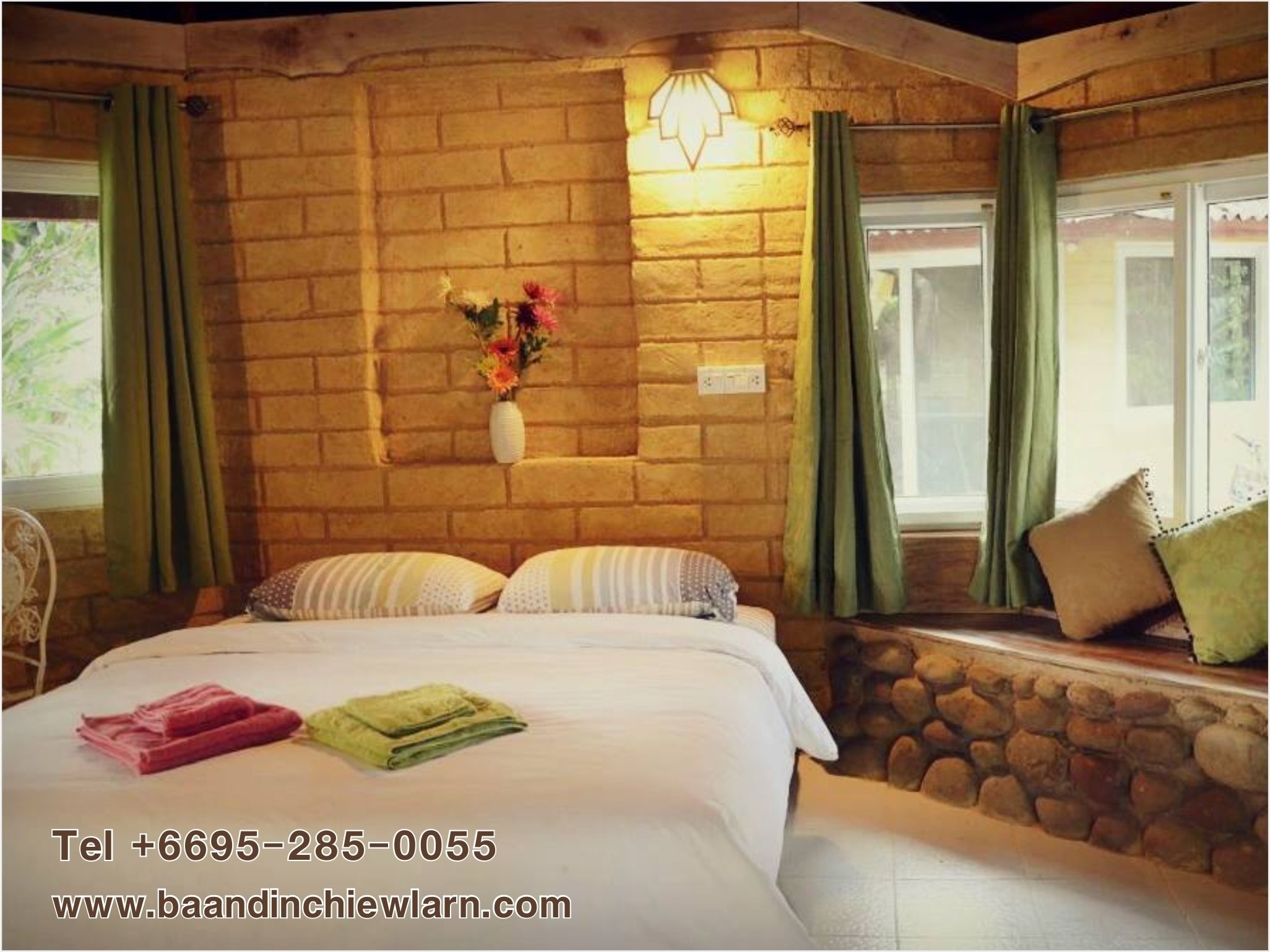 Una habitación con pared de ladrillo, cama con almohadas, un jarrón con flores y una ventana con cortinas.