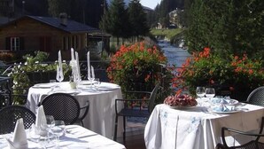 An outdoor dining area with tables set for a meal, surrounded by greenery and a view of a mountain path. - Hotel Baita Fiorita (Valfurva)