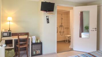 Une chambre d’hôtel comprenant un lit, un bureau avec une chaise, une télévision, une lampe, un four à micro-ondes, un petit réfrigérateur, une poubelle et un espace douche.