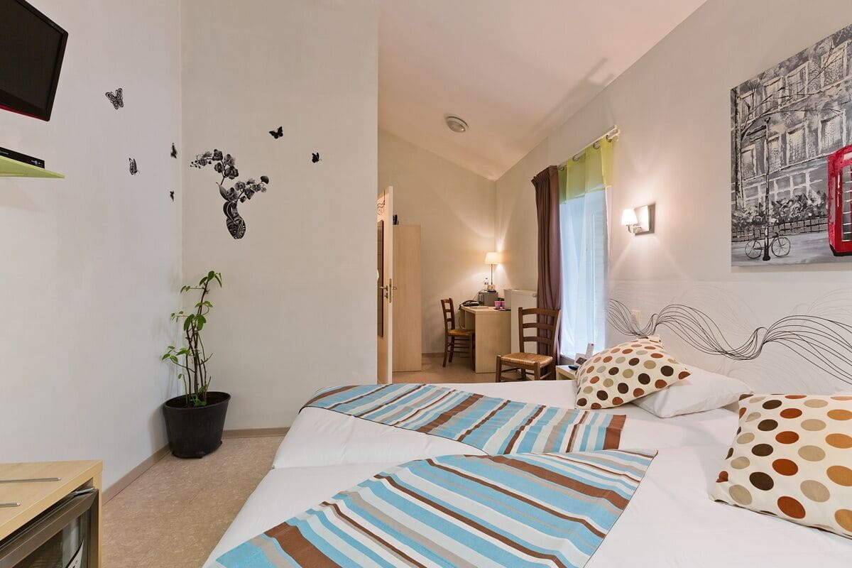 Een hotelkamer met een bed, een bureau, een stoel, een potplant en een aan de muur bevestigde televisie.