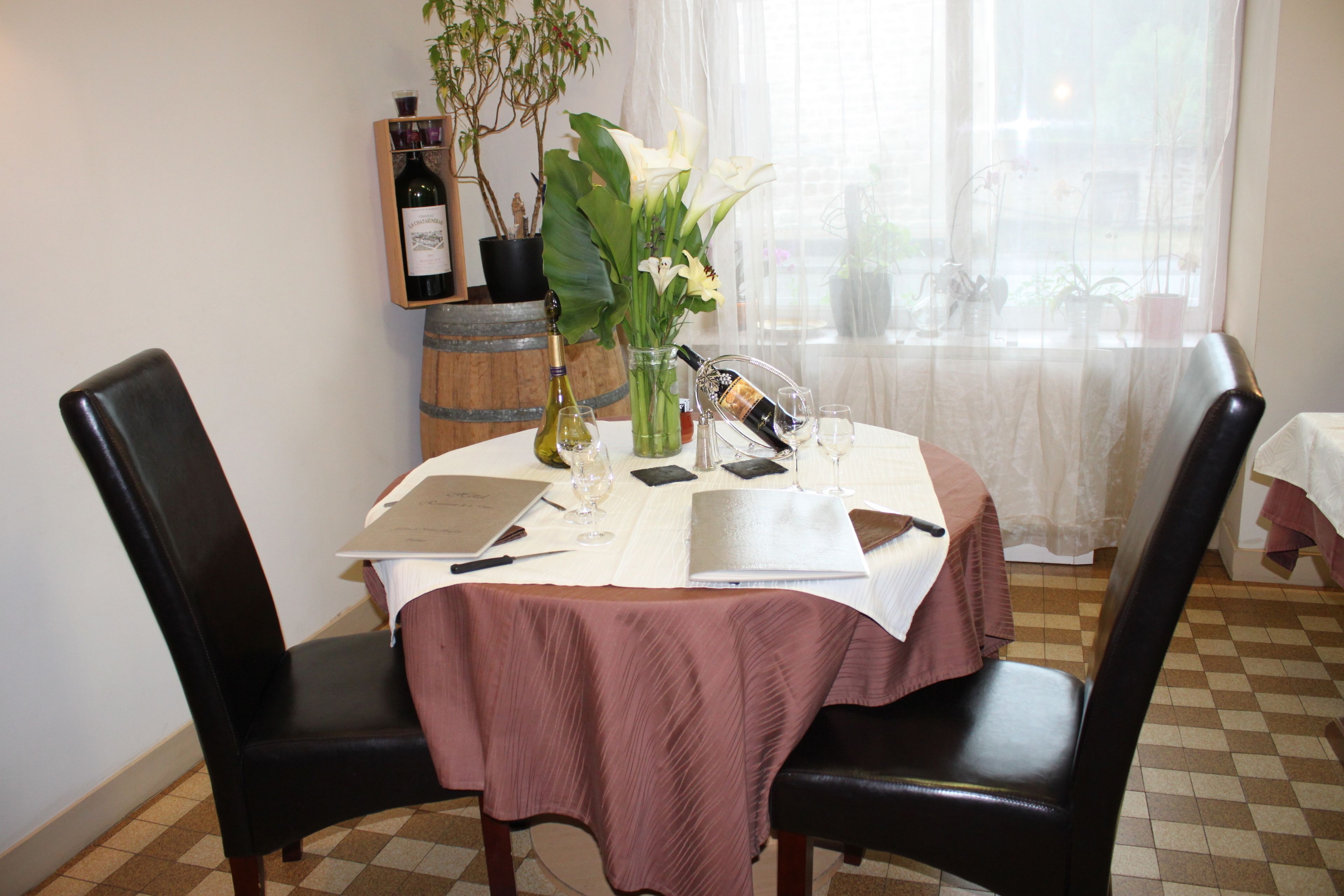 A dining table with a white tablecloth, two black chairs, a vase with white flowers, a bottle of wine, and a menu.
