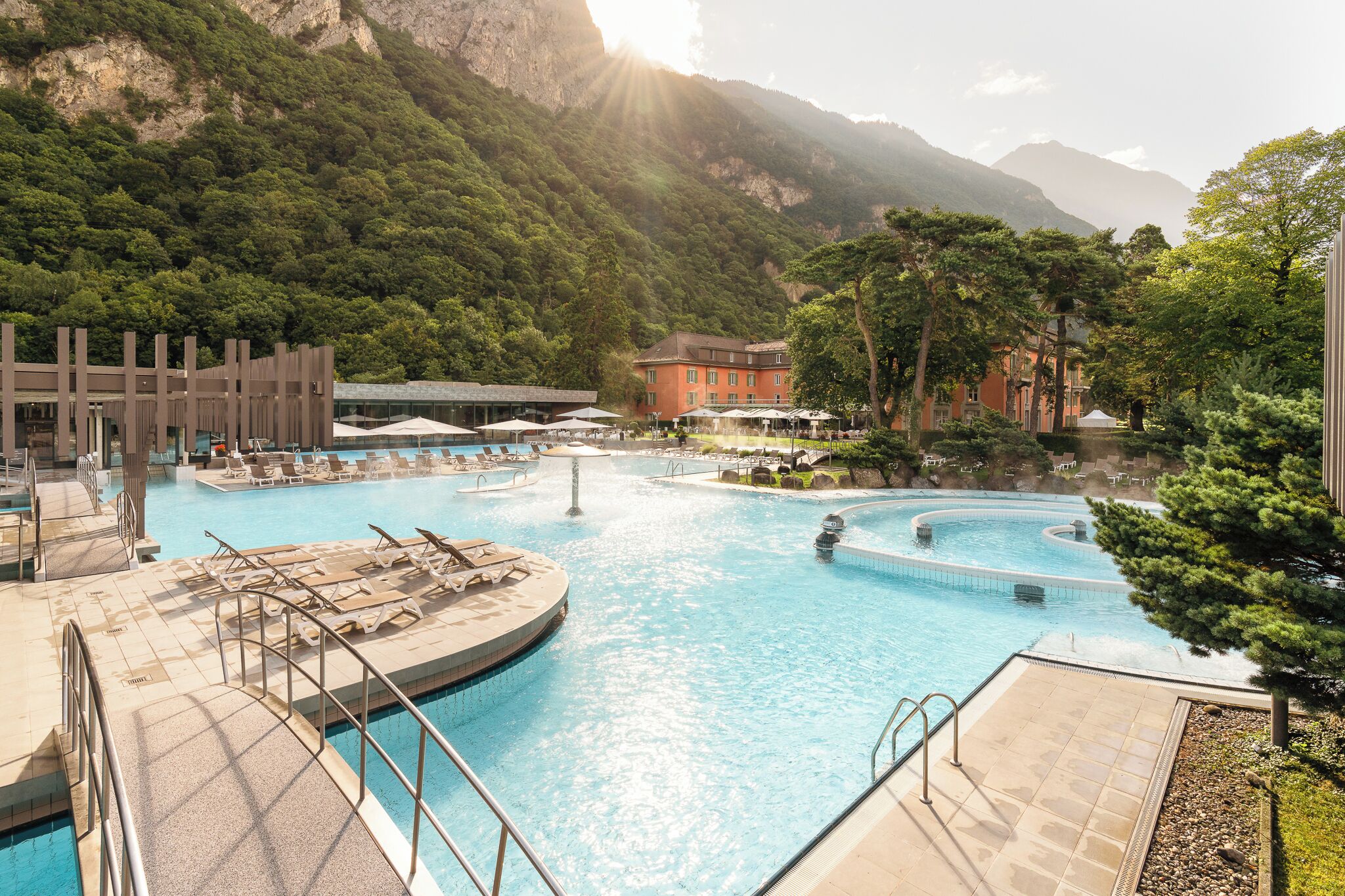 Zona de piscina con varias secciones, sillones de descanso y un edificio al fondo.