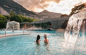 A pool with a waterfall feature, a covered seating area, and a mountain backdrop. - Grand Hôtel des Bains de Lavey (Lavey-Morcles)