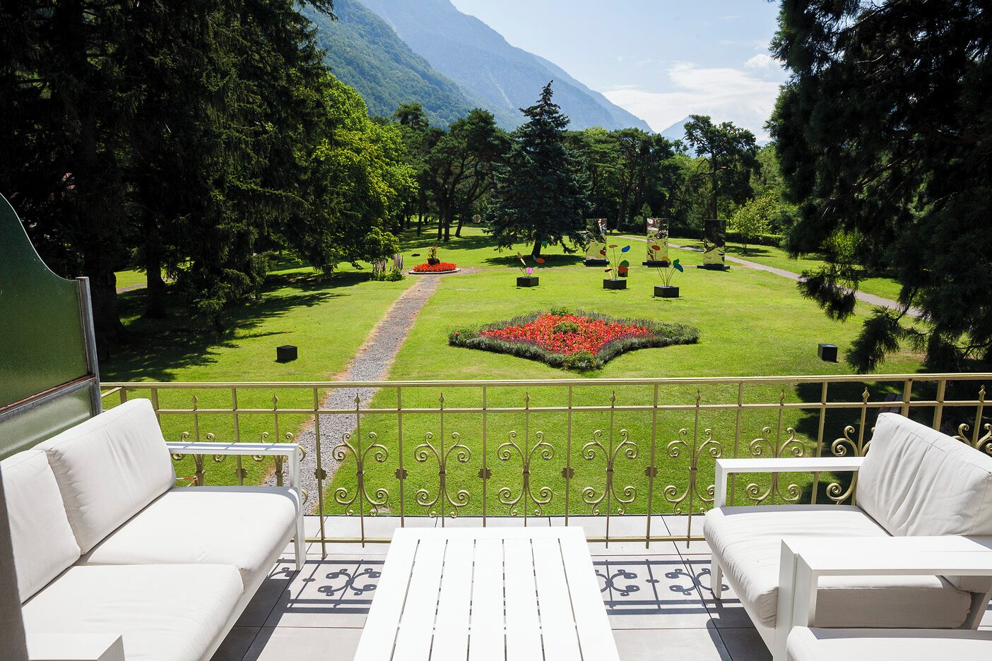 Un balcón con mobiliario exterior blanco, vista a un jardín cuidado con un parterre central, y un paisaje montañoso al fondo.