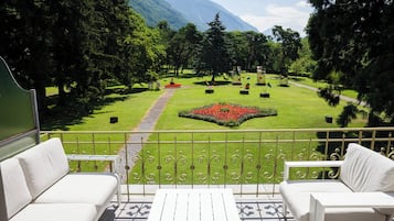 Un balcón con mobiliario exterior blanco, vista a un jardín cuidado con un parterre central, y un paisaje montañoso al fondo.