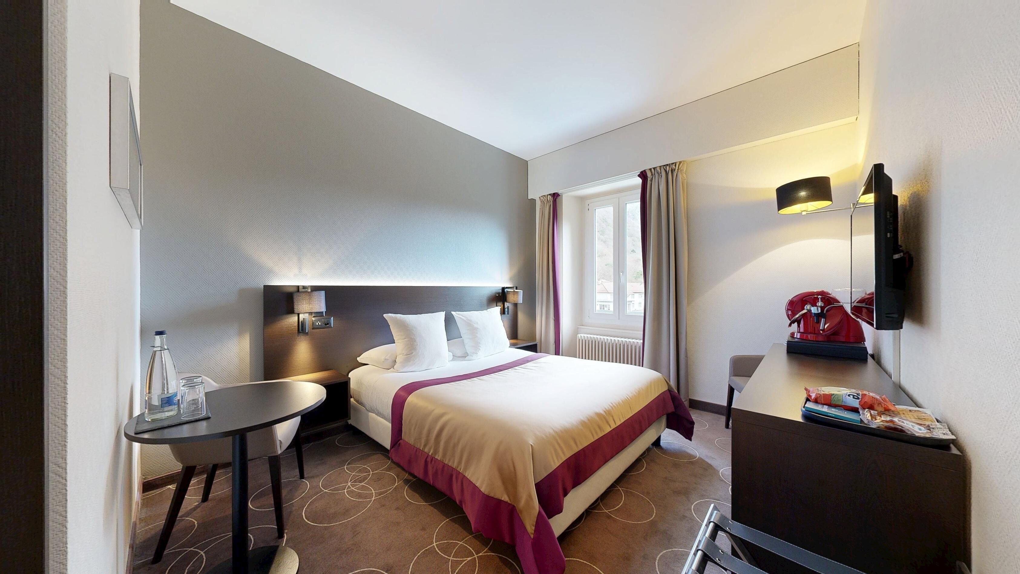 Ein Hotelzimmer mit Bett, Nachttisch, Lampe und einem kleinen Schreibtisch mit Stuhl.