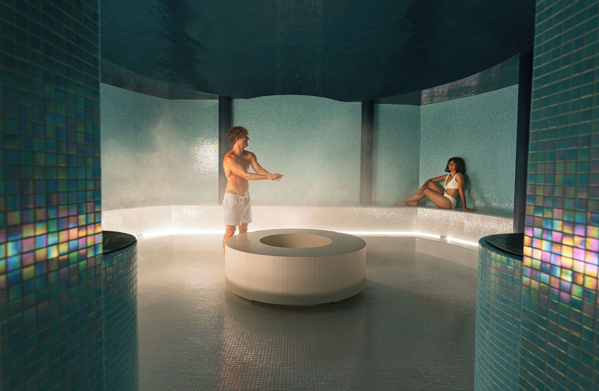 A modern indoor pool area with a central circular seating area, tiled walls, and a clear ceiling.